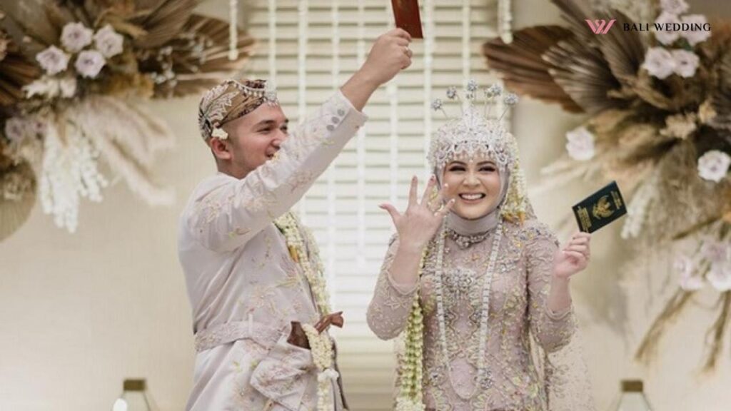 This image captures a joyful Indonesian wedding moment, with the bride and groom in elegant traditional attire. The groom raises a red booklet, while the bride proudly shows her marriage certificate and wedding ring.