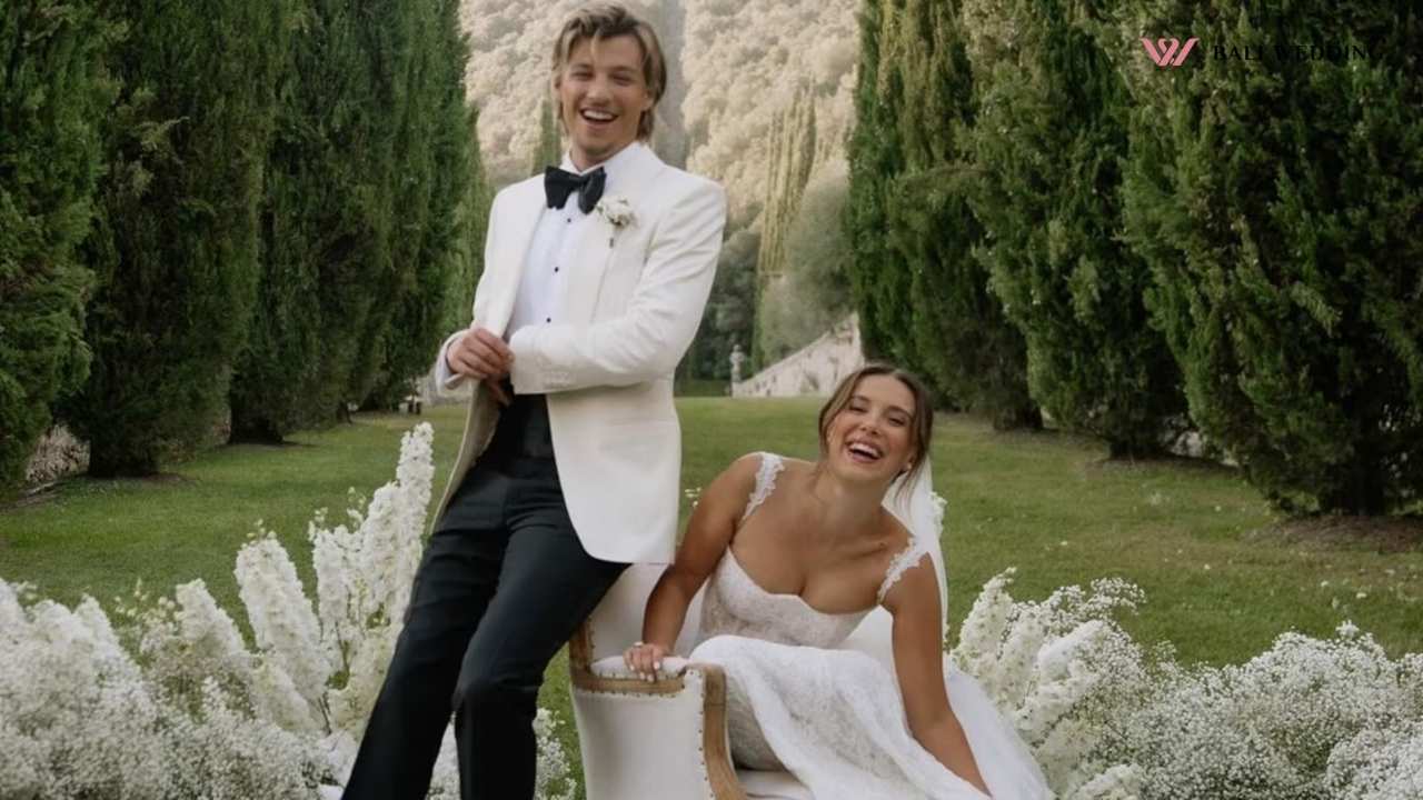 The groom and bride laughing in a green garden, the bride sitting in a white gown while the groom stands in a white suit.