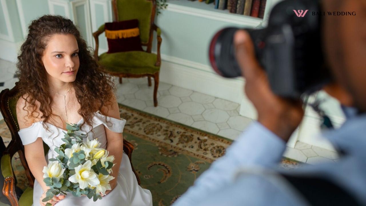 A photographer capturing a close-up shot of a bride sitting in an elegant room. The bride, wearing a simple white gown, holds a bouquet of white flowers and gazes off to the side.