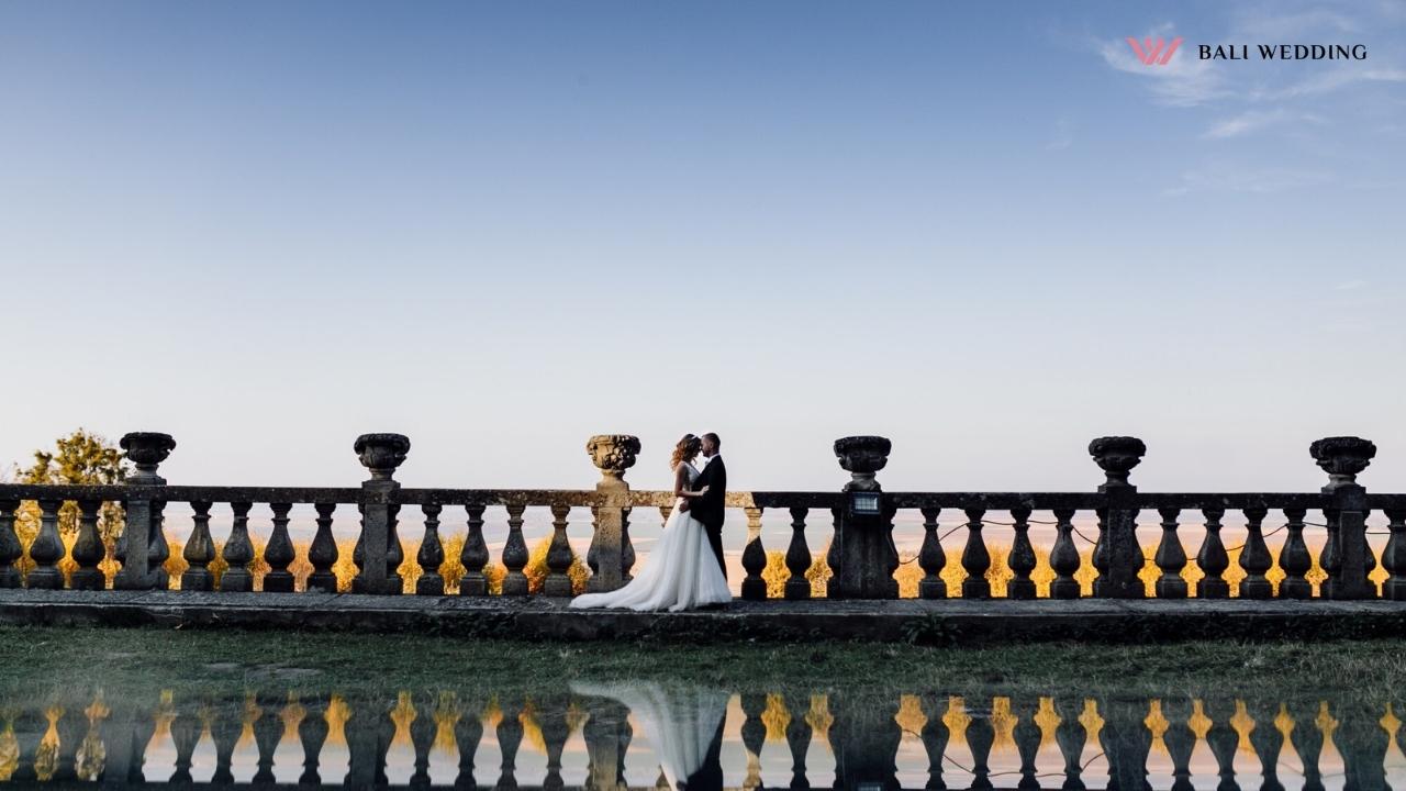 The bride and groom standing on a terrace with a beautiful view, their reflection visible in the water in front of them.