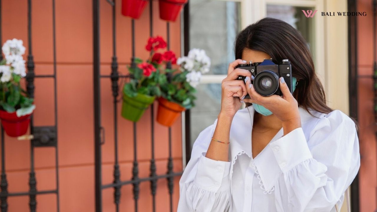 A woman holding a camera up to her face, ready to take a picture. She is standing outdoors, with a backdrop of flower pots attached to a wall.