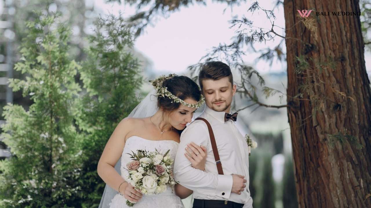 A bride and groom standing close to each other in a serene outdoor setting, surrounded by lush green trees. The bride holds a bouquet of roses and wears a floral headband, while the groom looks down at her affectionately.