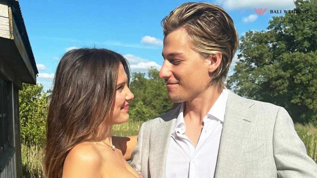 Millie and Jake are smiling at each other outdoors, with trees and a bright blue sky in the background.