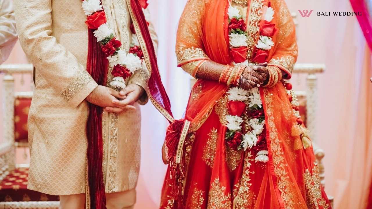 A bride and groom dressed in traditional Indian wedding attire, adorned with floral garlands, standing side by side, symbolizing a ceremonial moment of unity.